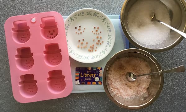 silicone snowman mould, rock salt pieces in the shape of eyes, nose and mouth, plus bowls full of a bath salt mixture consisting of epsom salt and pink himalayan rock salt
