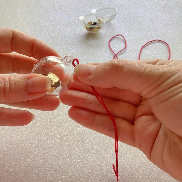a pair of hands threading a looped piece of red thread through a clear bauble